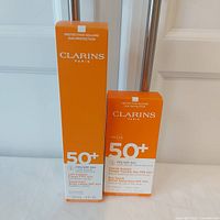 Clarins SPF 50+ sunscreen body lotion 150ml and dry touch facial sunscreen 50ml in orange and white boxes, standing side by side.