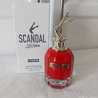 Front view of perfume bottle with box showing red bottle with gold cap shaped like legs and white plain box.