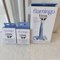 Overview photo showing one Flamingo pubic razor kit box with light blue razor handle, two boxes of replacement cartridges each containing 4 blades.