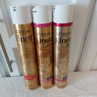 Three L'Oreal Elnett hairspray cans in gold and white with pink top accents, standing upright.