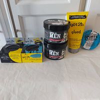 Photo showing all six hair styling products: 2 Dippity-do men texture pomade jars stacked, Schwarzkopf got2b sculpt paste and spiking wax in boxes, Schwarzkopf got2b spiking glue tube, Johnny's Chop Shop styling fibre tin, all on flat surface.