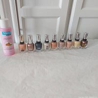 Front view of 9 Sally Hansen nail polish bottles in various nude, pink, bronze shades with silver caps and one pink bottle of Vitry nail polish remover with almond oil, all placed on a flat white surface against a door background.