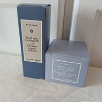 Photo of two boxed Naturium skincare products including BHA Liquid Exfoliant 2% (120ml) in a blue box and Purple Ginseng Cleansing Balm (88ml) in a purple box.