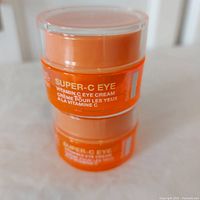 Two stacked bright orange jars of StriVectin Super-C Eye Vitamin C Eye Cream with clear lids on a white textured surface.