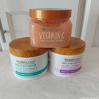 Three body scrub jars shown: Tree Hut Vitamin C Shea Sugar Scrub on top, two Raw Sugar Salt Polish Daily Micro Scrubs below in different scents and white jars with wood-textured lids.