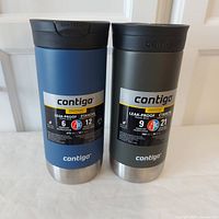 Two Contigo travel mugs side by side, blue and grey/olive with labels showing capacity and insulation times.