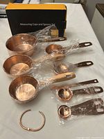 Copper-finish stainless steel measuring cups and spoons laid out with original packaging on table, each piece partially wrapped in plastic, alongside a copper metal ring for holding them together.