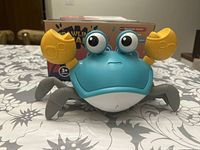 Front view of the blue and gray plastic crab toy with yellow claws and large eyes on stalks, placed on a patterned tablecloth with its box shown behind it.