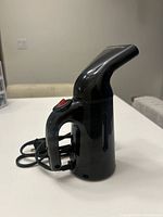 Photo of black handheld electric garment steamer showing side profile with steam nozzle angled upward, power switch visible on handle, power cord wrapped behind device.