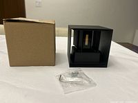 Black metal cuboid-shaped wall mounted light fixture next to cardboard box and plastic bag with screws on white surface.