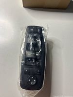 Front view of the black plastic car window switch showing multiple buttons and labels for window and mirror controls, inside a plastic bag.