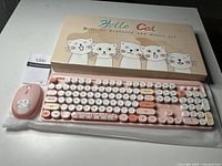 Photo showing the pink wireless keyboard with round keys and cartoon cat designs, next to matching pink wireless mouse and original box with cartoon cat faces.