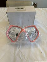 Clear plastic safety goggles with red trim in plastic wrapping, positioned in front of a white cardboard box on a table.