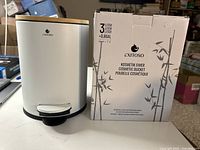 Front view of the cosmetic bucket with its box. White body, bamboo lid, foot pedal, and brand logo visible.