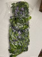 5 bundles of green and purple artificial flower garlands bundled in plastic wrap, showing leafy green vines with purple fabric flowers.