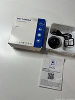 Image showing unopened box labeled WiFi Camera with model and certification icons, camera body, USB cable, mounting frame, and YsxLite app instruction sheet.