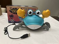 Photo showing blue and yellow plastic crab toy with big eyes, legs, claws, and USB charging cable next to original retail box.