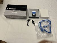 Linksys NPV network storage system with original box, white inner box, black USB cable, and blue Ethernet cable in plastic laid out on white surface.
