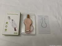 Photo showing the pink WA04S wireless watch charger in plastic blister packaging next to its manual and the retail box showing product features.