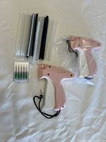 Two pink plastic tagging guns, one still wrapped in plastic, laid out on a white surface with multiple strips of plastic fasteners and small plastic cases containing green needle attachments.