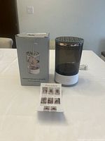 Photo shows ultrasonic makeup brush cleaner device with translucent grey body, white base with power button, and stainless steel perforated lid, alongside its box and instruction sheet.