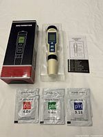 Digital water quality tester in molded plastic packaging with retail box showing front image and control buttons. Three pH buffer powder packets labeled 4.00, 6.86, and 9.18, plus instruction sheet included.