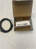 Wired industrial sensor inside open cardboard box alongside black cable coiled with exposed wire ends, clear plastic mounting plate and metal screw also visible.