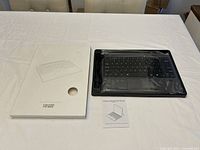 White original box with keyboard image and text indicating Surface Pro wireless keyboard, alongside black wireless keyboard in plastic wrap, USB cable, and user manual on a white surface.