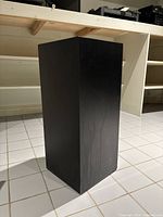 Front angle view of black melamine display pedestal showing rectangular shape and surface finish.