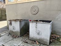 Two large square outdoor planters with noticeable dirt and weathering, placed on a paved surface near a wall.