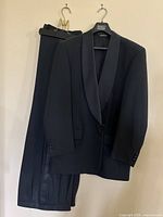 Black tuxedo jacket and trousers hanging on hangers, showing full front view of suit components.