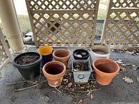 Twelve garden pots arranged on wooden stand outdoors under a trellis. Includes various sizes, materials such as terracotta, plastic, and metal. Shows wear and dirt from outdoor use.