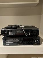 Two stacked Sony units; top is Betamax player/recorder with remotes on top, bottom a VCR unit, all black with multiple buttons and input ports visible.