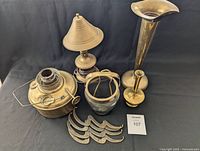 Group photo showing all brass items together on a black backdrop: tall trumpet-shaped vase, brass lamp base with small lampshade, brass ice bucket, small candle holder, and four ornate hooks