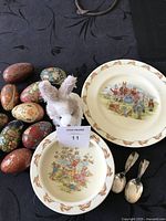 Top view of two Bunnykins ceramic plate and bowl, set of 11 decorative Easter eggs, TY Hobsy bunny plush, and two silver-plated spoons arranged on fabric