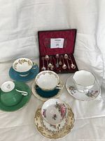 Five bone china teacup and saucer sets displayed with box of silver plate teaspoons in back