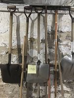 Photo showing the full set of garden tools hanging against a white stone wall including shovels, rakes, and forks.
