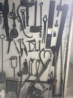 Wide view of multiple vintage metal tools hanging on pegboard, including tongs, hooks, clamp, and hand-forged tools.