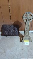 Photo of both the concrete Celtic cross garden statue and the cast iron wood stove door side by side against a wooden panel wall on concrete floor.
