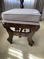 Foot stool front view showing beige padded cushion and carved solid wood base on castors.
