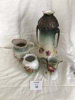 Four porcelain items on a white fabric background: tall Royal Vienna vase with handles and brown top, a floral pitcher, a small floral vase, and a FERN England decorated vase.