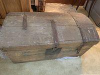 Full view of rustic wooden trunk with hinged lid, curved top, and metal straps showing rust, placed on carpeted floor.
