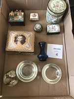 Top down photo of mixed collection of small trinket and pill boxes, small glass vessels and decorative items inside a cardboard box.