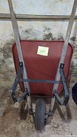 Front view of wheelbarrow showing red metal barrel and wooden handles resting on floor against wall.