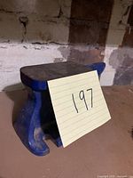 Side view showing blue anvil with a note '197' placed on it, on a dusty surface with brick wall background.