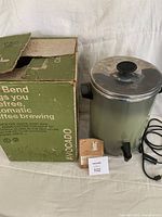 Vintage avocado-colored automatic coffee maker with stainless steel lid, side handle, and black knob. Positioned next to original green box and instruction booklet on white fabric surface.