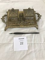 Full view of the vintage brass double inkwell stand with dip pen placed in front, showing ornate design, handles, and hinged lids closed.