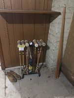 Wide shot showing entire croquet set on metal stand with several wooden balls, two wooden pool cues leaning against wall and pair of wooden shoe forms on floor.