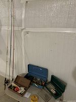 Photo showing three tackle boxes with open lids revealing various fishing lures, hooks, bobbers, and fishing accessories inside. Boxes include one metallic blue and two plastic (brown and green).