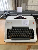 Front view of the white Olympia manual typewriter showing black keys and gold logo, paper loaded in platen.